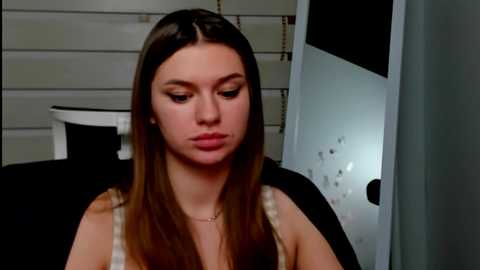 Media: Video of a young Caucasian woman with straight brown hair, wearing a white top, sitting in a black chair, looking contemplative, in a dimly lit room with white horizontal blinds and a dark door.