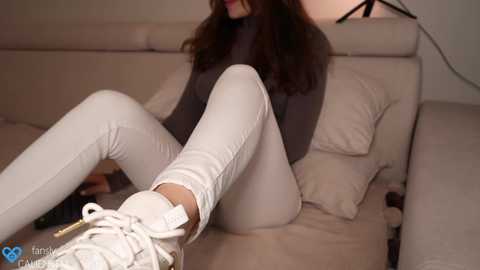 Media: Video of a woman with long brown hair, wearing a grey turtleneck and tight white pants, sitting on a beige couch, legs crossed, with white sneakers.