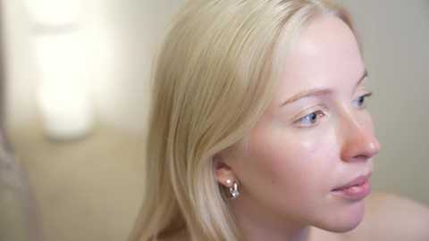 Media: A close-up video of a fair-skinned, blonde woman with straight hair and light makeup, wearing silver hoop earrings. She gazes to the side in a softly lit room.