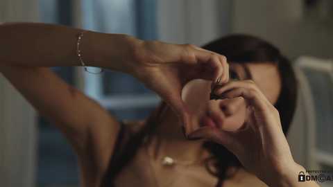 Media: A video shows a woman with medium brown skin, long dark hair, and small breasts, forming a heart shape with her fingers near her face. She wears a delicate bracelet and necklace. The background is blurred, featuring a window.