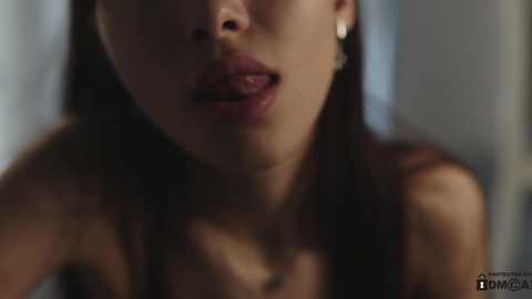 Media: Close-up video of a woman with light brown skin and long black hair, wearing a sleeveless top, her lips slightly parted, set against a blurred, dimly-lit background.