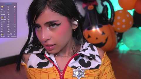 Media: Video of a young Latina woman with long black hair, wearing a cow-print jacket, black eyeliner, and a Halloween pumpkin balloon in the background.