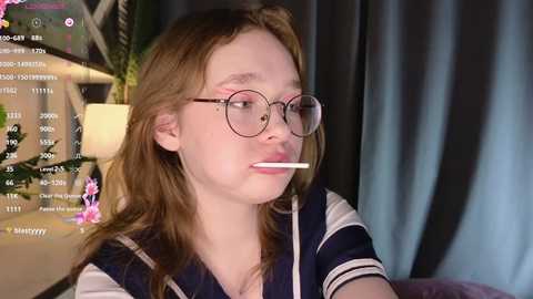 Media: A young, fair-skinned girl with long, wavy brown hair, wearing round glasses, ponders with a cigarette in her mouth. Background shows a blurred, dark curtain and a green plant.