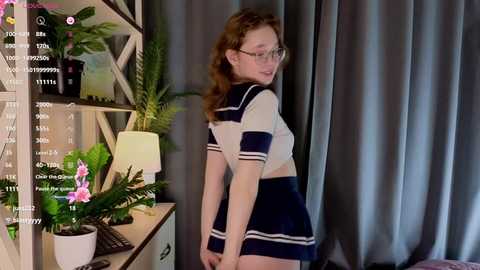 Media: Video of a fair-skinned woman with long red hair and glasses, wearing a navy blue sailor schoolgirl outfit, standing in a room with grey curtains, plants, and a white shelf.