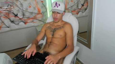 Media: Video of a young, light-skinned, shirtless man with a tattoo on his chest, wearing a white hat with \"As\" logo, sitting at a computer in a modern room with abstract art on the wall.