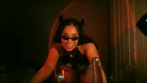 Media: A video depicts a woman with long black hair, wearing devil horns and sunglasses, leaning seductively over a glass table, bathed in dim red light.