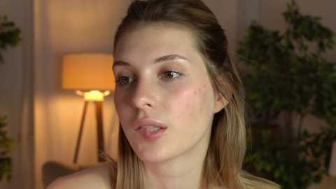 Media: A video of a young Caucasian woman with light skin, brown eyes, and straight blonde hair, wearing a sleeveless top, indoors near a lamp and green plants.