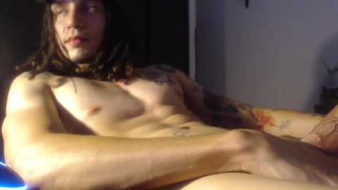 Media: Video of a nude, fair-skinned man with long brown hair, tattoos, and a flaccid penis. He is lying on a bed with dim lighting, creating shadows on his body.