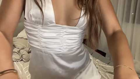 Media: Video of a woman in a white, ruched, sleeveless dress, with long brown hair, indoors, on a bed with beige sheets, wearing a gold bracelet on her left wrist.