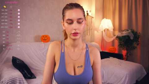 Media: Video of a fair-skinned woman with light brown hair in a ponytail, wearing a blue tank top, sitting on a bed with a Halloween pumpkin. Background includes a lamp, plants, and a calendar.