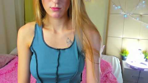 Media: Video of a young woman with long blonde hair, wearing a blue tank top, standing in a cozy bedroom with a pink blanket, white headboard, and fairy lights.