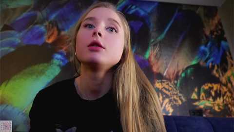 Media: Video of a young blonde woman with fair skin and blue eyes, wearing a black t-shirt, looking contemplative against a colorful abstract background.