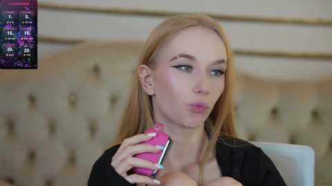 Media: Video of a fair-skinned, blonde woman with straight hair, wearing a black top, blowing into a pink phone. She has light makeup with winged eyeliner. Background features a beige tufted headboard and a digital screen displaying a score.