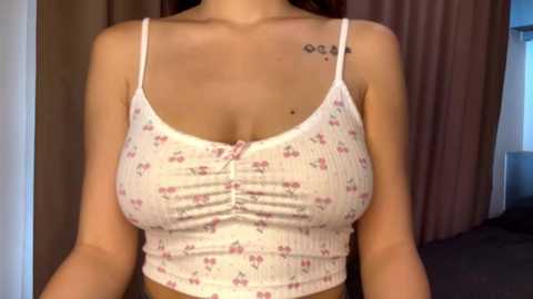 Media: Video of a light-skinned woman with medium-sized breasts, wearing a white ribbed tank top with small floral patterns, revealing a tattoo on her left shoulder. Background shows beige curtains and a blue wall.