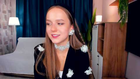 Media: Video of a young woman with long, straight brown hair, wearing a black dress with silver butterfly embroidery, silver necklace, and matching earrings, smiling in a modern living room with a white sofa, wooden cabinet, and blue curtains.