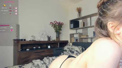 Media: Video of a partially nude woman with light skin, wearing a black thong, lying on a bed with a patterned duvet. A wooden dresser with a vase of flowers and a TV screen displaying a fitness app are in the background.