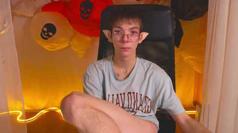 Media: Video of a pale-skinned, young man with glasses, pointy elf ears, and short dark hair, wearing a \"Heavy D\" T-shirt, seated in a black chair, surrounded by Halloween-themed decorations in a dimly lit room.