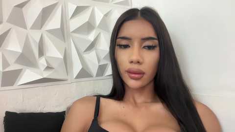 Media: Video of a young woman with long black hair, wearing a black bra, standing against a white wall with geometric white patterns. She has a tan complexion and full lips.