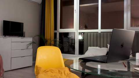 Media: Video of a modern, minimalist bedroom with a yellow chair, white dresser, and open glass doors. A closed laptop and open MacBook are on a glass table, and a bed with white sheets is visible through the doors.