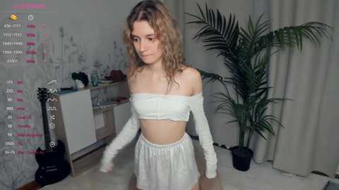 Media: Video of a slender, fair-skinned woman with wavy blonde hair, wearing a white off-shoulder crop top and matching high-waisted shorts, standing in a cozy, modern room with a tall plant and a guitar.