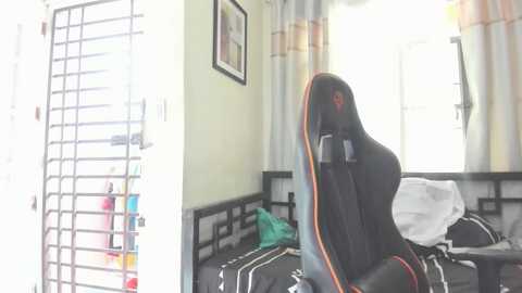 Media: Video of a modern gaming chair with orange accents, positioned in a bright bedroom with light green walls and a window with white curtains. The room is cluttered with bedding and clothes on the bed.