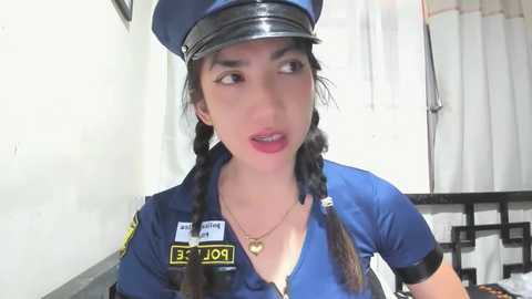 Media: Video of a young Asian woman with dark hair in braids, wearing a blue police uniform, including a hat, badge, and heart necklace, standing indoors near a white wall and black railing.