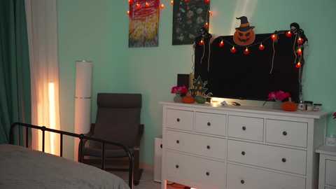 Media: A video of a cozy bedroom with pastel green walls, a white dresser adorned with Halloween decorations, a black-framed mirror, and a gray chair.