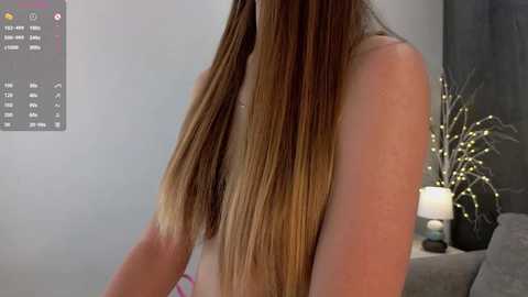 Media: Video of a topless woman with long, straight, blonde hair, standing indoors. Background features a calendar, a candlelit floral arrangement, and a gray couch.