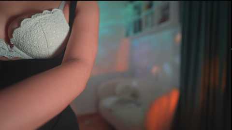 Media: A close-up video of a woman in a white lace bra, with blurred background of a dimly lit room featuring soft, warm lighting and a hint of greenery.