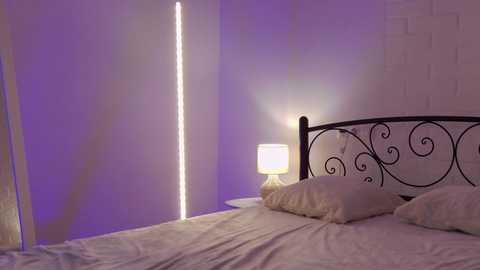 Media: A video of a minimalist bedroom with a white brick wall, a black wrought iron bed with a floral headboard, and a bedside table with a white lamp casting soft light.