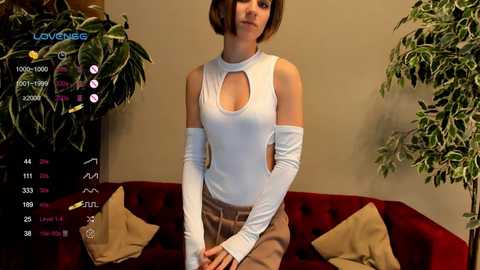 Media: Video of a woman with short brown hair, wearing a white, long-sleeved top with cutouts, gray pants, and elbow-length gloves, standing in front of a red velvet couch with beige pillows.