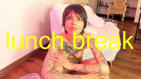 Media: Video of a tattooed woman sitting naked on a wooden floor, looking contemplative. Background shows a bed, chair, and shelves in a dimly lit room. \"Lunch break\" is overlaid in yellow text.