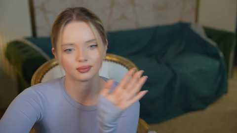 Media: Video of a young white woman with fair skin, light brown hair tied back, wearing a light blue sweater, making a hand gesture in a softly lit, vintage-style room with a dark green sofa and floral wallpaper.