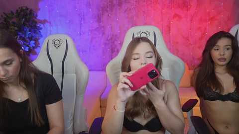 Media: Video of three young women seated in a gaming chair, with a red phone in the foreground. One wears a black bra, another a black top, and the third a white top. Background features a purple and red lighting effect.