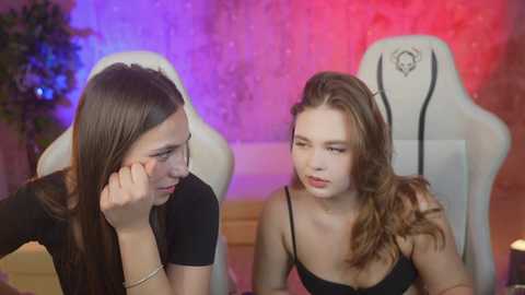 Media: Video of two young women, one with long brown hair, the other with wavy brown hair, sitting side-by-side in a gaming chair, in a dimly lit room with purple and red lighting.