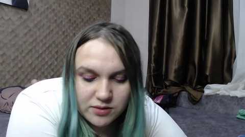 Media: Video of a young woman with light skin and shoulder-length teal hair, wearing purple eyeshadow, a white top, and sitting on a bed with a textured brown wall behind her.