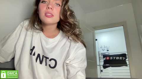 Media: A video of a young woman with wavy brown hair, wearing a white \"MTNT\" sweatshirt, standing indoors near a white door.