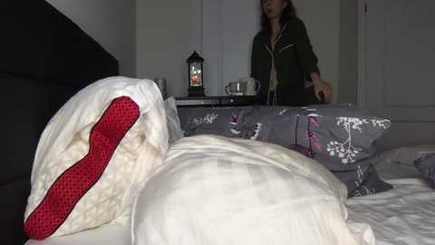 Media: Video of a woman with shoulder-length brown hair, in a dark green jacket, standing beside a bed with a white comforter and a pillow with a red heart pattern. The background features a closed white door and a nightstand with a lantern.