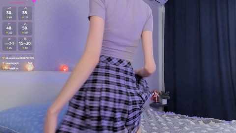 Media: A video of a slender woman in a light gray t-shirt and black-and-white plaid skirt, captured from behind while dancing on a bed.