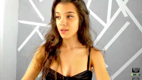 Media: Video of a young woman with long, wavy brown hair, fair skin, and dark eyes, wearing a black lace bra, standing against a geometric grey wall.