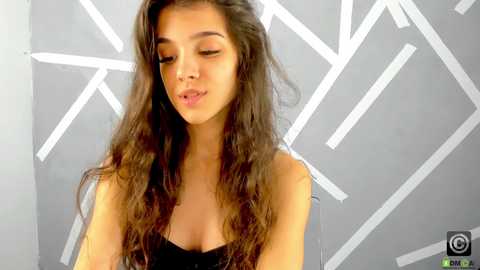 Media: Video of a young woman with long, wavy brown hair, wearing a black strapless top, against a modern geometric grey wall with white lines.