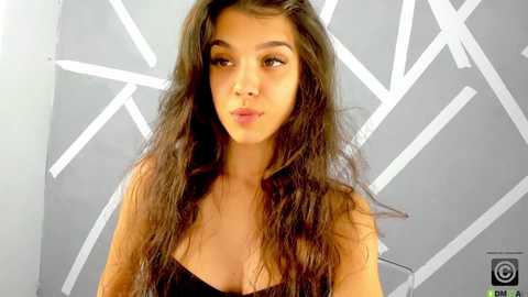 Media: Video of a young woman with long, wavy, dark brown hair, wearing a black strapless dress, standing against a geometric, white-patterned grey background. She has a light olive skin tone and a neutral expression.