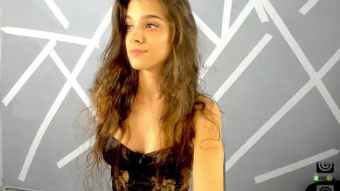 Media: Video of a young woman with long, wavy brown hair, wearing a black lace top, standing against a geometrically patterned gray wall. She has a light skin tone and a contemplative expression.