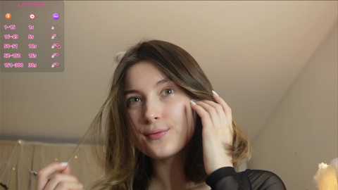 Media: Video of a young Caucasian woman with shoulder-length, wavy brown hair, light skin, and a slight smile. She's indoors, adjusting her hair. Background includes a beige wall and a digital clock displaying a date and time.