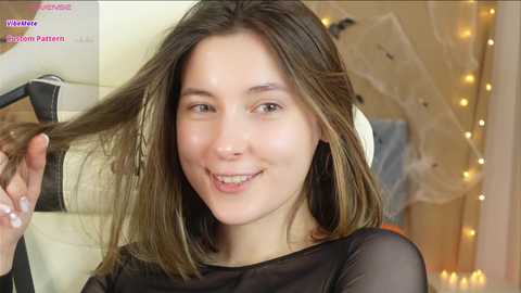 Media: Video of a young woman with light skin and straight brown hair, smiling while holding a strand of hair. She wears a sheer black top. Background features a soft, ambient lighting and white fabric.