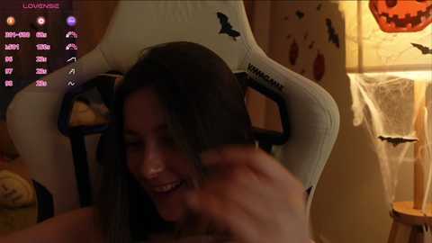 Media: A video captures a young woman with long, dark hair, smiling widely, seated in a white gaming chair, with Halloween decorations in the background.