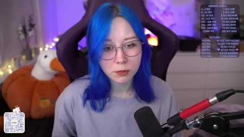 Media: Video of a pale-skinned woman with blue hair and glasses, wearing a grey shirt, recording a video with a microphone on a black gaming chair in a dimly lit room with a stuffed duck and a purple wall.