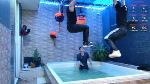 Video of two men in black workout gear hanging from a fitness trapeze above an indoor swimming pool, surrounded by blue and orange equipment.