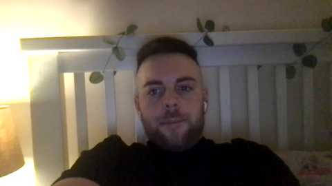 Media: Video of a bearded man with short, dark hair, wearing a black shirt, sitting on a white crib with green vines and lights, in a dimly lit room.