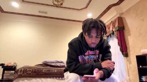 Media: Video of a young Black man with dreadlocks in a cozy bedroom, wearing a black hoodie with \"Hustle\" text, sitting on a bed, playing with a pink phone.